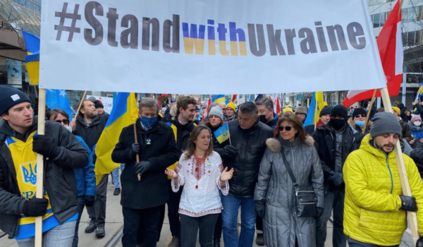 A group of people marching down a street wih a white banner being held by two people on each side. The banner reads out, Stand with Ukraine.