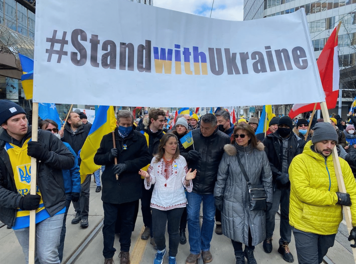 A group of people marching down a street wih a white banner being held by two people on each side. The banner reads out, Stand with Ukraine.