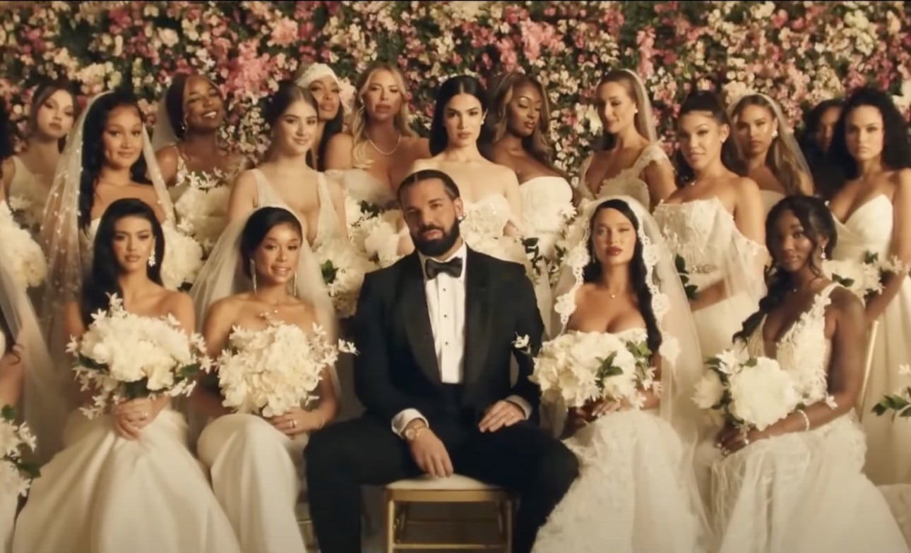 Drake in a black tux, sitting with a crowd of women in wedding dresses